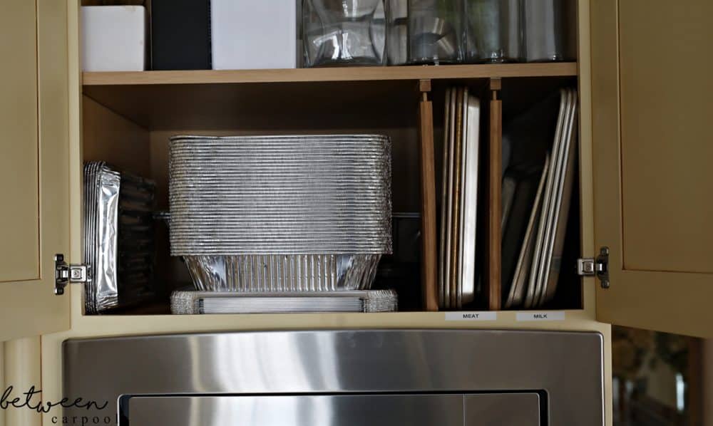Top cabinet with drawer dividers installed vertically. Metal sheet pans stored between the dividers. A stack of 9x13 foil pans and covers on the shelf