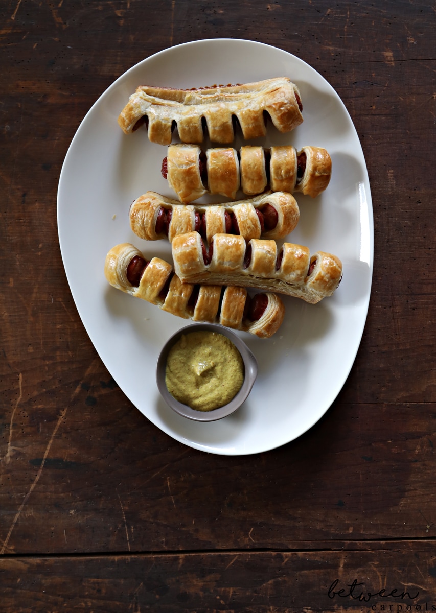 pull apart hot dogs, baked, on a white oval tray with mustard in a stainless condiment bowl