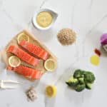 Slices of salmon and lemon on unbleached parchment paper. Mayonnaise, seasoning, lemon, brown rice, and special sauce in a dip bowl. (a top view image of all ingredients for Salmon, Rice and Broccoli dinner)