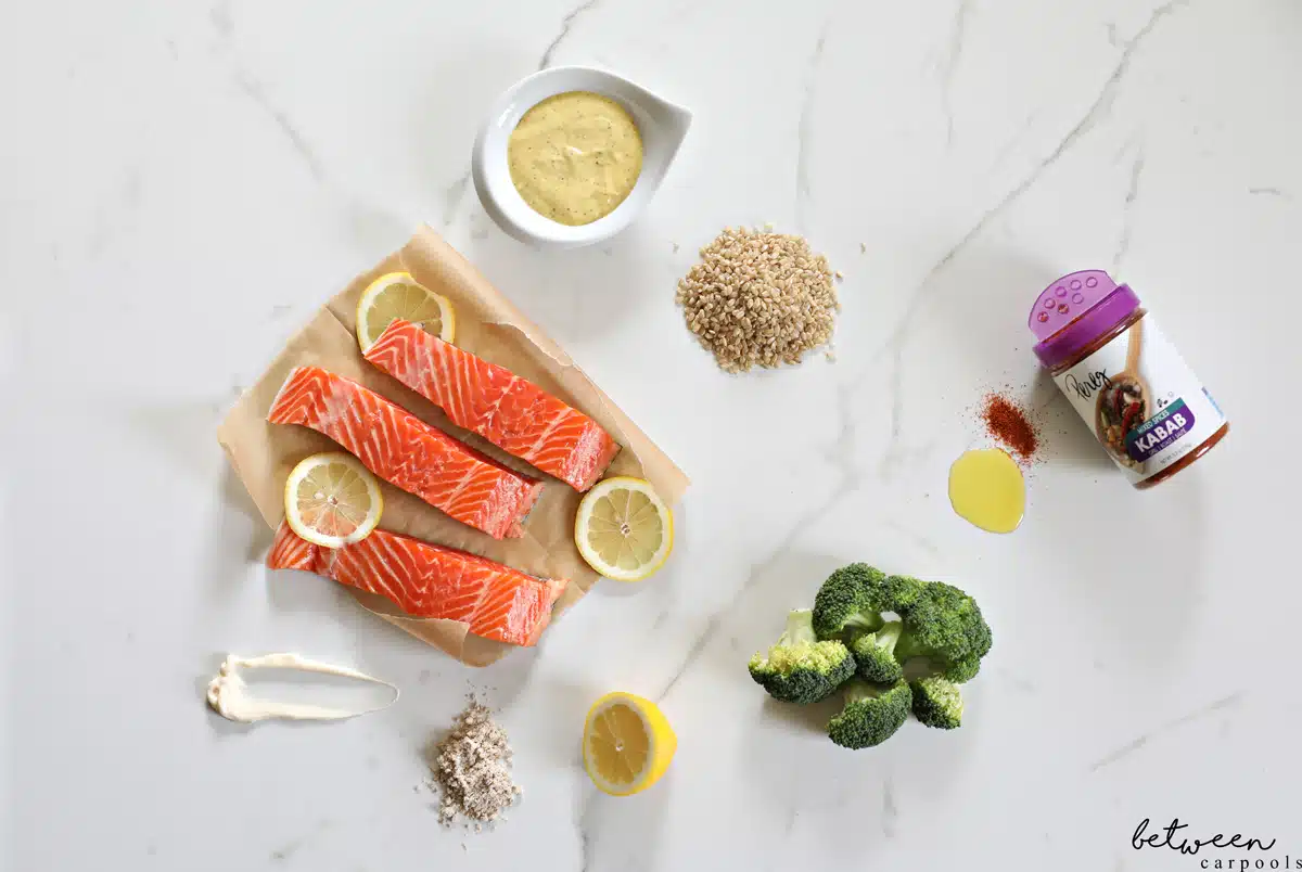 Slices of salmon and lemon on unbleached parchment paper. Mayonnaise, seasoning, lemon, brown rice, and special sauce in a dip bowl. (a top view image of all ingredients for Salmon, Rice and Broccoli dinner)