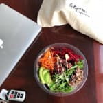 An assembled poke bowl in a plastic salad bowl on a table with car keys and a MacBook.