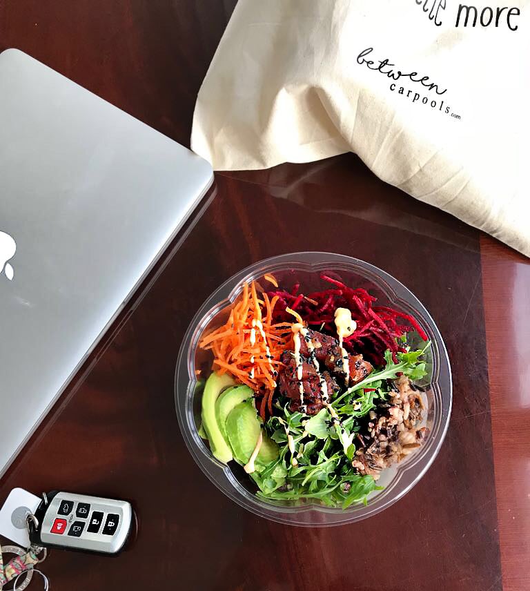 An assembled poke bowl in a plastic salad bowl on a table with car keys and a MacBook.