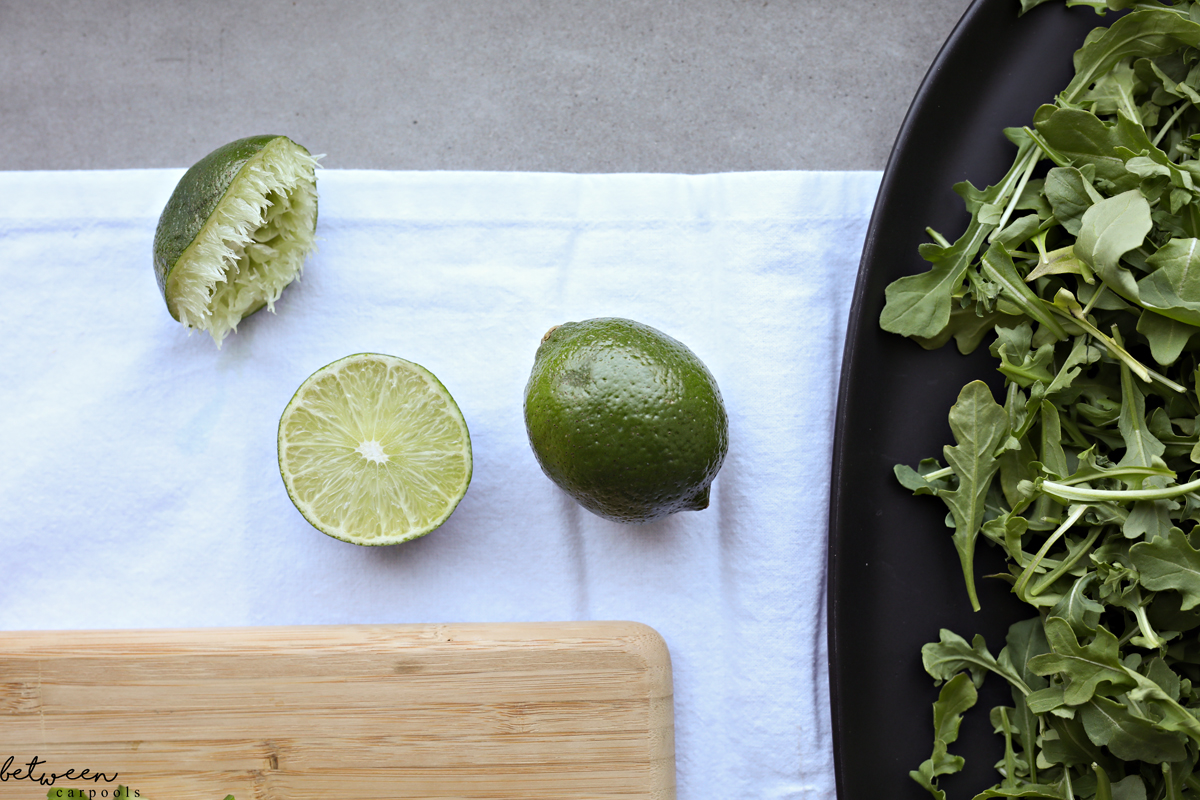 Arugula on a black oval platter. One lime, one half lime, one half squeezed lime and a wooden cutting board.
