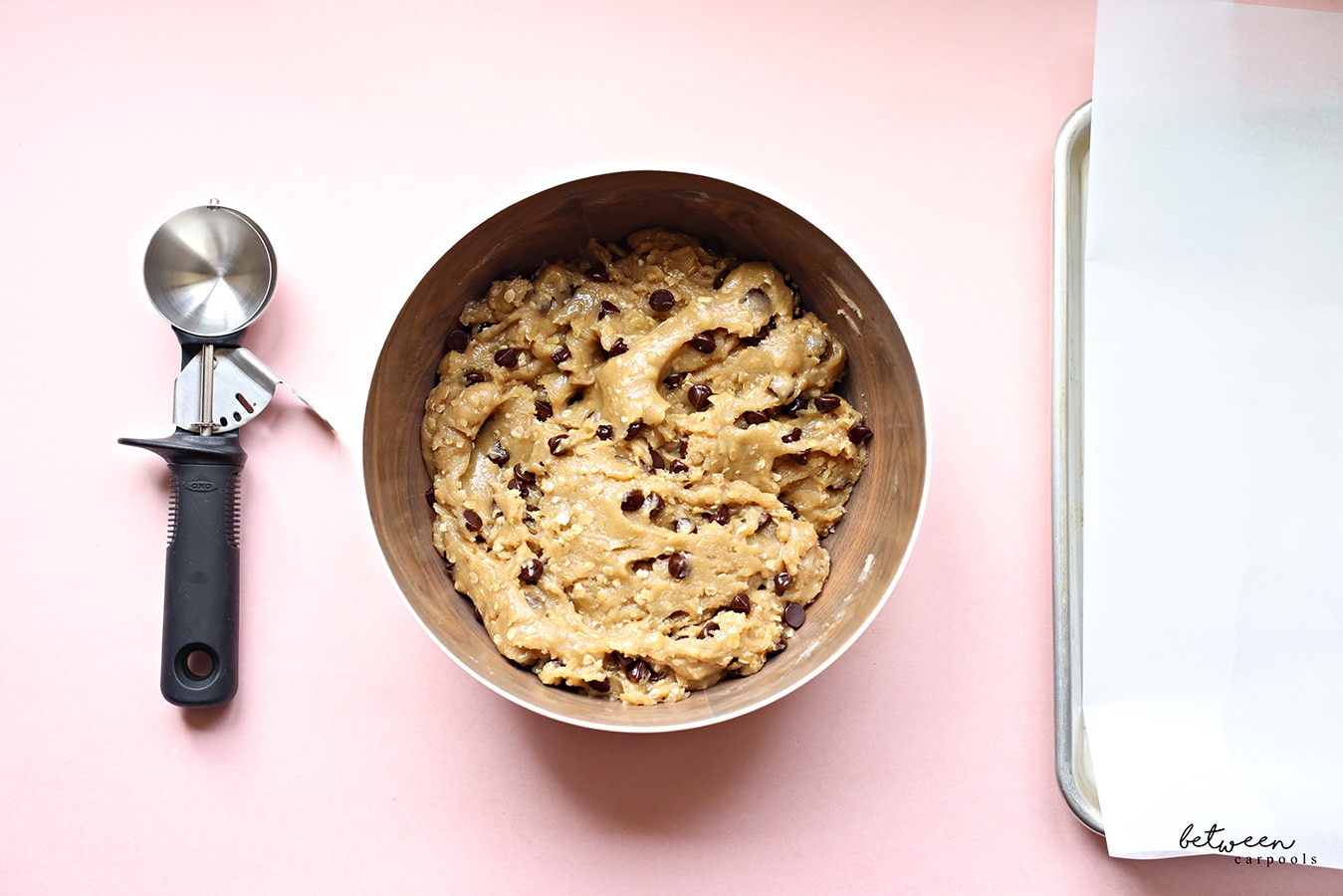 Ice cream scoop, chocolate chip cookie dough in a metal bowl and a metal sheet pan lined with parchment paper.