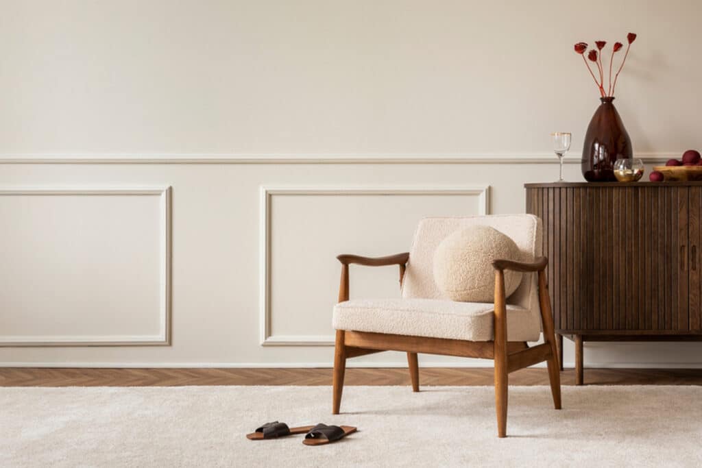 Aesthetic composition of living room interior with boucle armchair, vase with dried flowers, round pillow, wooden sideboard, beige rug and personal accessories.