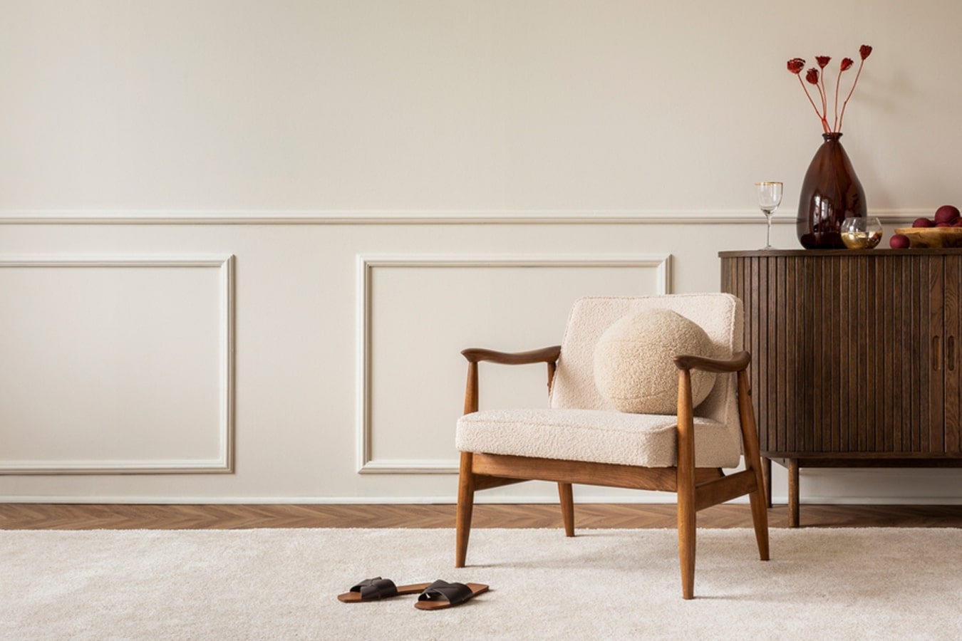 Aesthetic composition of living room interior with boucle armchair, vase with dried flowers, round pillow, wooden sideboard, beige rug and personal accessories.