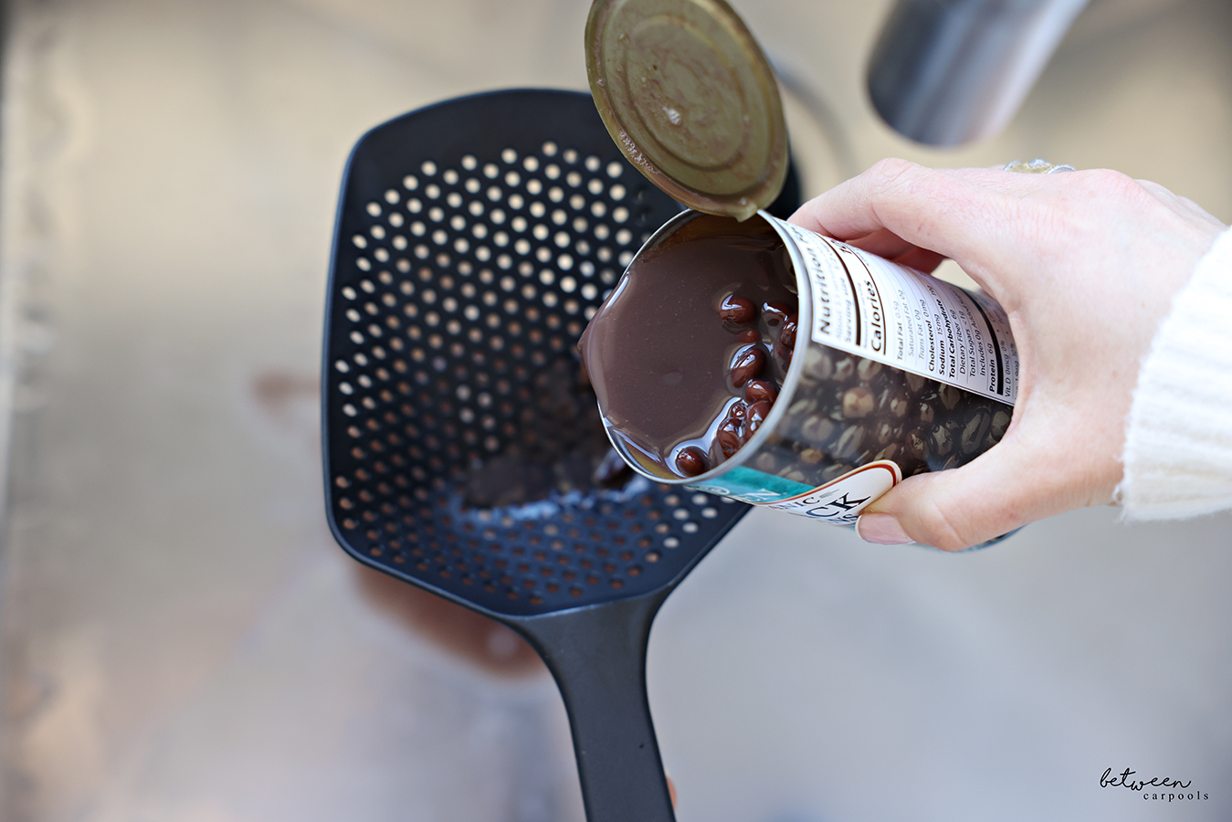 Pouring a 15 oz can of Eden Organic Black Beans into a black plastic scoop colander over the sink.