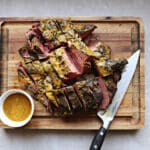 Sliced baked Navel Pastrami and brown sugar/mustard sauce over it, on a cutting board. Knife and extra sauce in condiment dish.