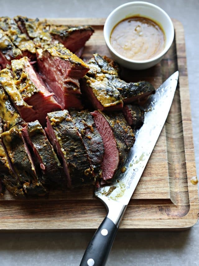 Sliced baked Navel Pastrami and brown sugar/mustard sauce over it, on a cutting board. Knife and extra sauce in condiment dish.