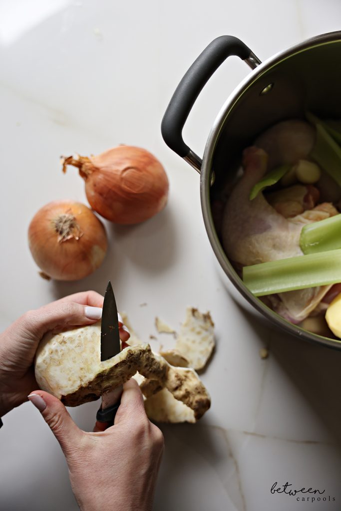 Peeling a celery knob with a knife. Chicken, golden potatoes and celery stalks in an 8 quart pot. Onions nearby.