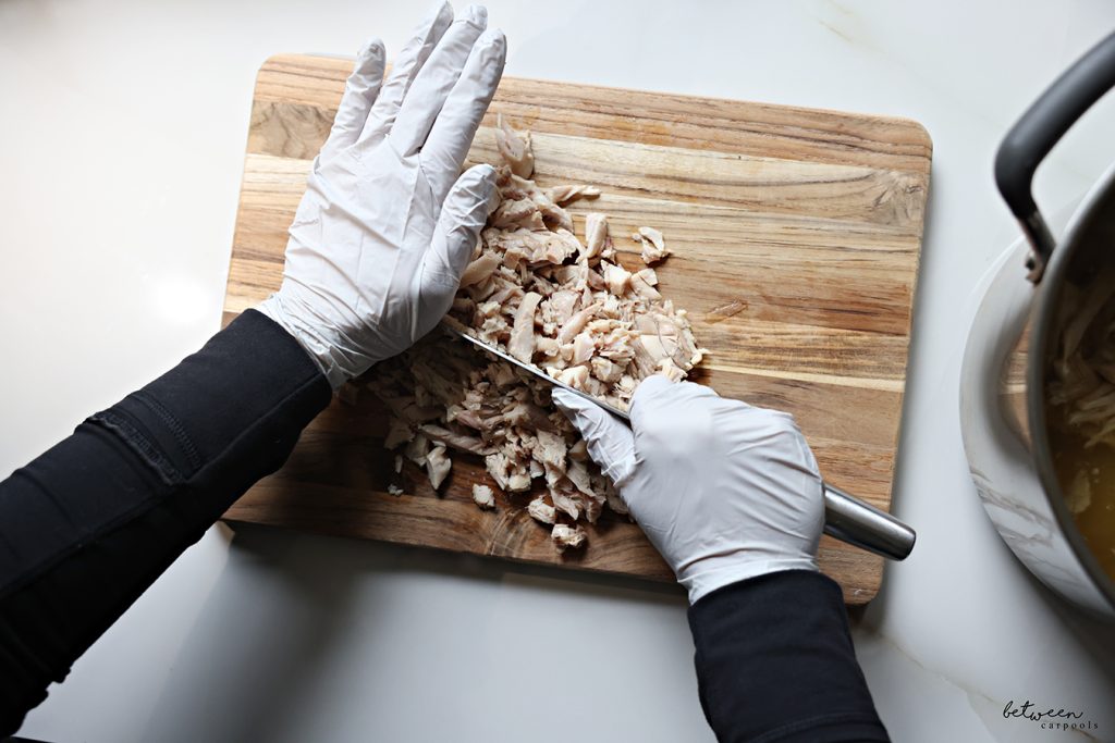 Chopping chicken bottoms on a wooden cutting board with gloved hands and a hast knife.