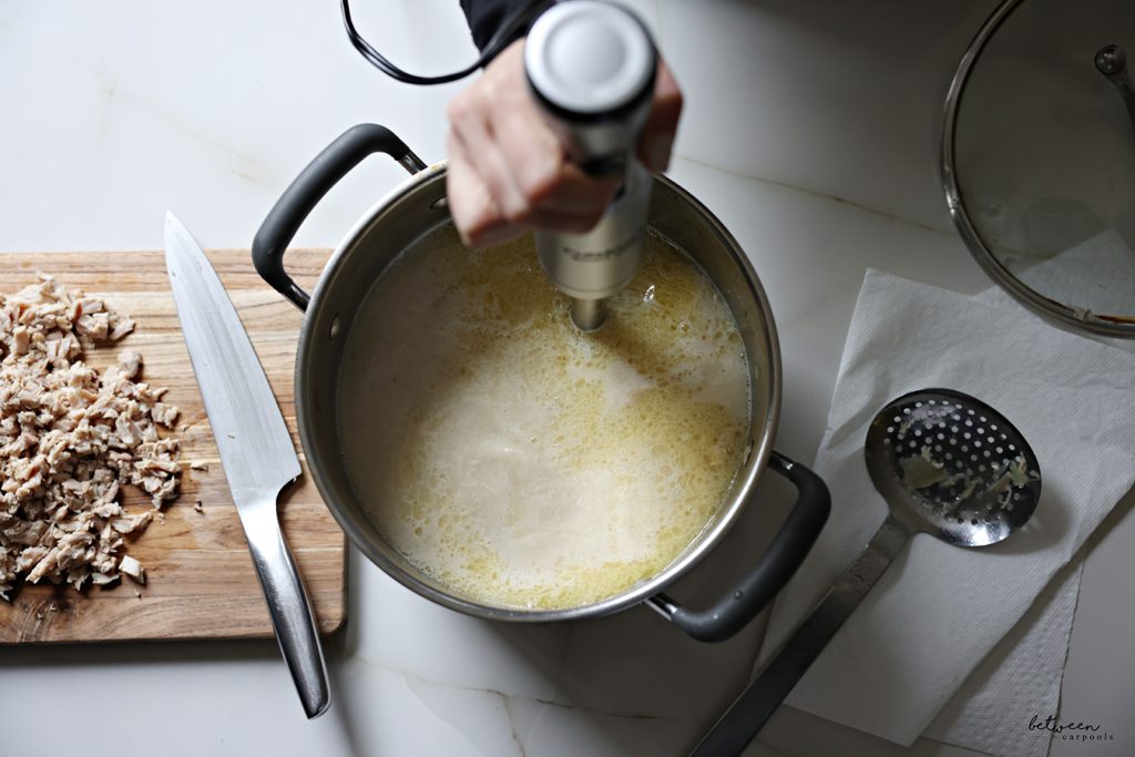 Blending chicken broth with its vegetables and chicken tops using an immersion blender in an 8 quart pot.