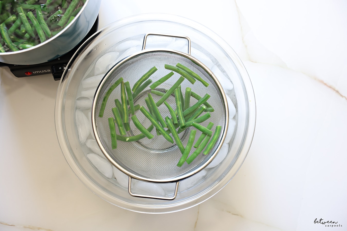 Blanched green beans in a colander in a bowl of water with ice cubes.