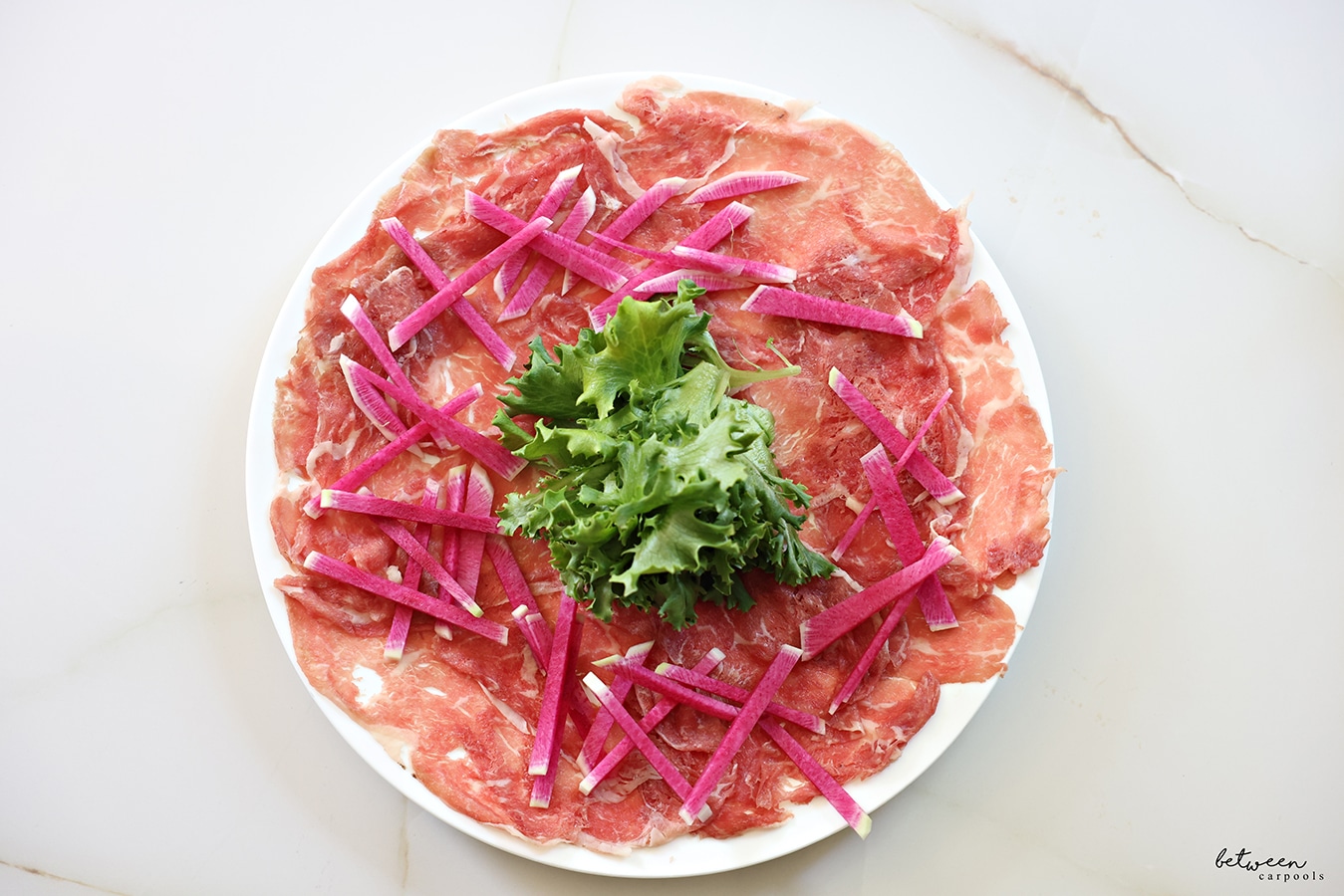 a plate of carpaccio meat, greens in the center and sliced watermelon radish around