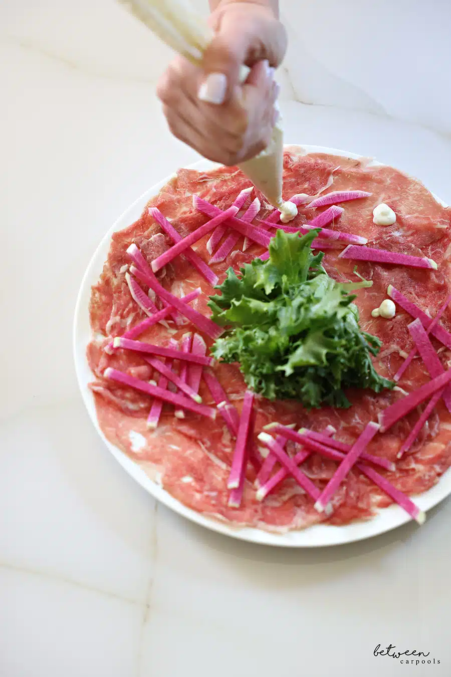 adding garlic aioli to a plate of carpaccio meat with greens in the center and sliced watermelon radish around