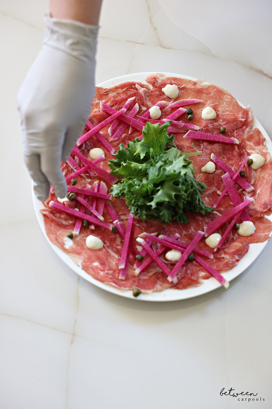 aioli dotted all around on the plate of carpaccio meatwith greens in the center and sliced watermelon radish around