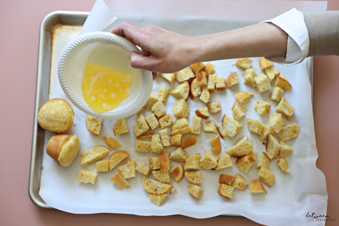 pouring melted butter over cubed corn muffins on a parchment paper lined sheet pan tray