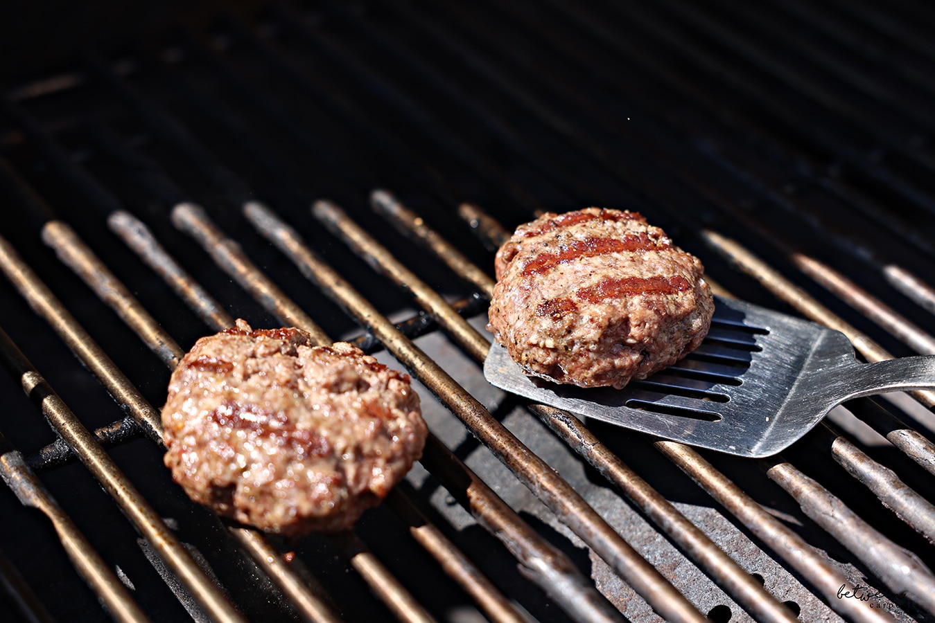 2 grilled burgers on an outdoor grill. Spatula under one of them