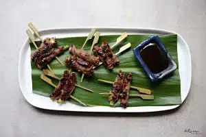 grilled roll ups on a banana leaf on a white oval platter. teriyaki sauce in a dish on the leaf