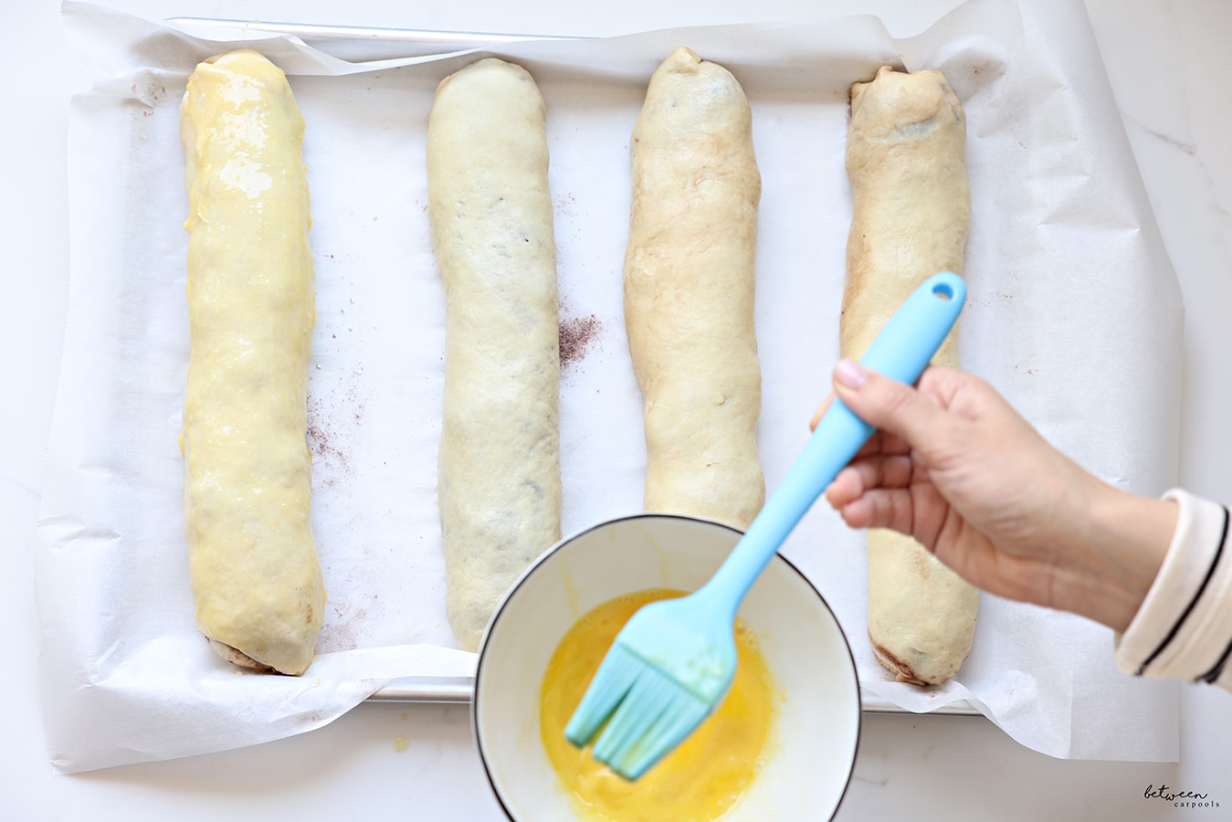 4 rolls of kokosh cake on one baking sheet. Holding a silicone egg brush and beaten eggs in a bowl