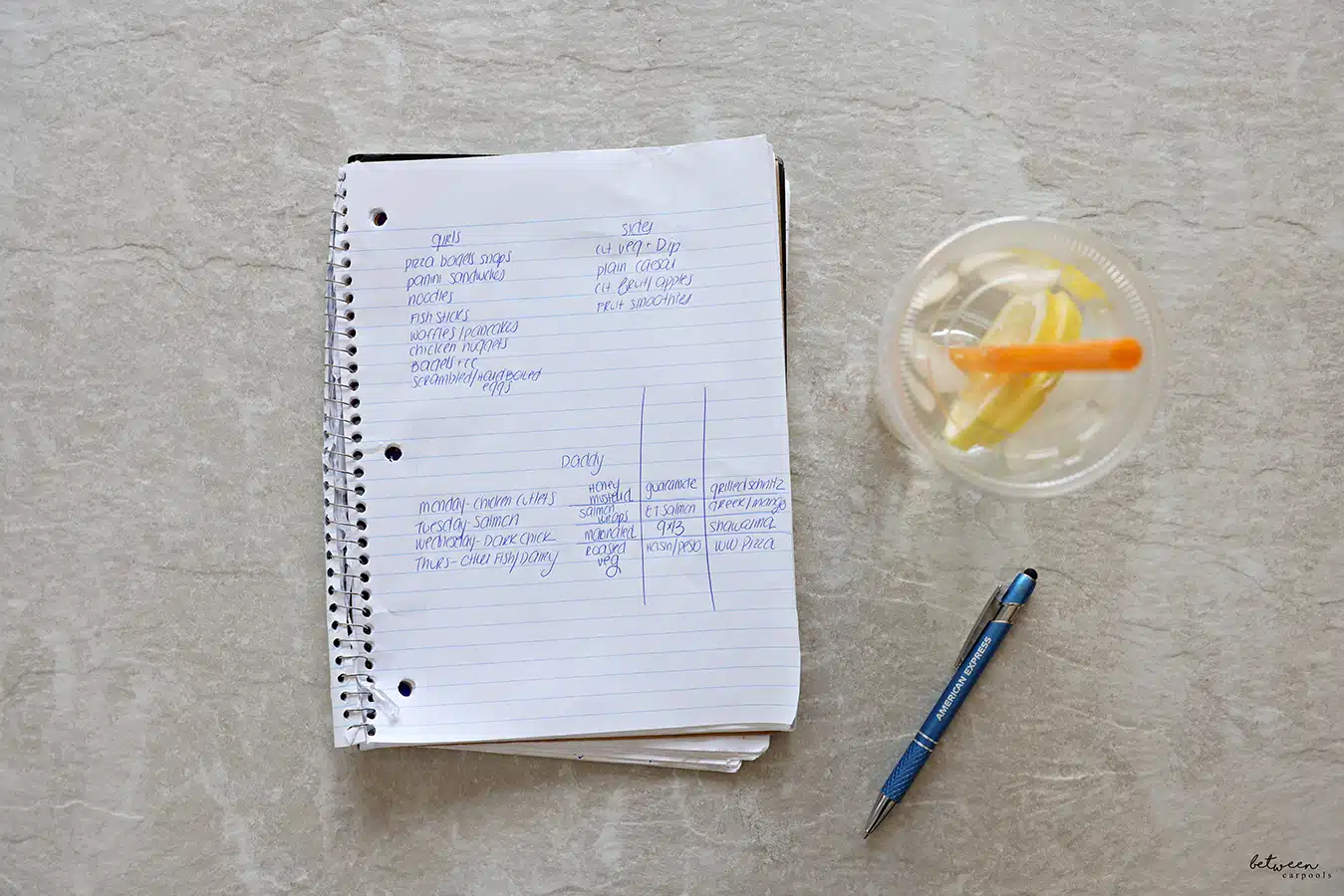Handwritten menu plan in a spiral notebook. Pen and lemon water nearby