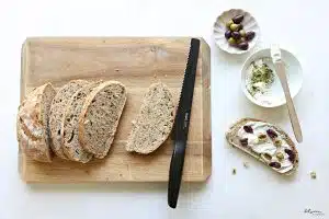 No Knead Artisanal Bread - sliced on a cutting board with a black hast bread knife - one slice topped with soft cheese, herbs and olives. soft cheese plus herbs and olives in bowls