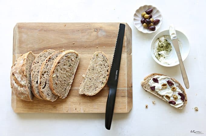 No Knead Artisanal Bread - sliced on a cutting board with a black hast bread knife - one slice topped with soft cheese, herbs and olives. soft cheese plus herbs and olives in bowls