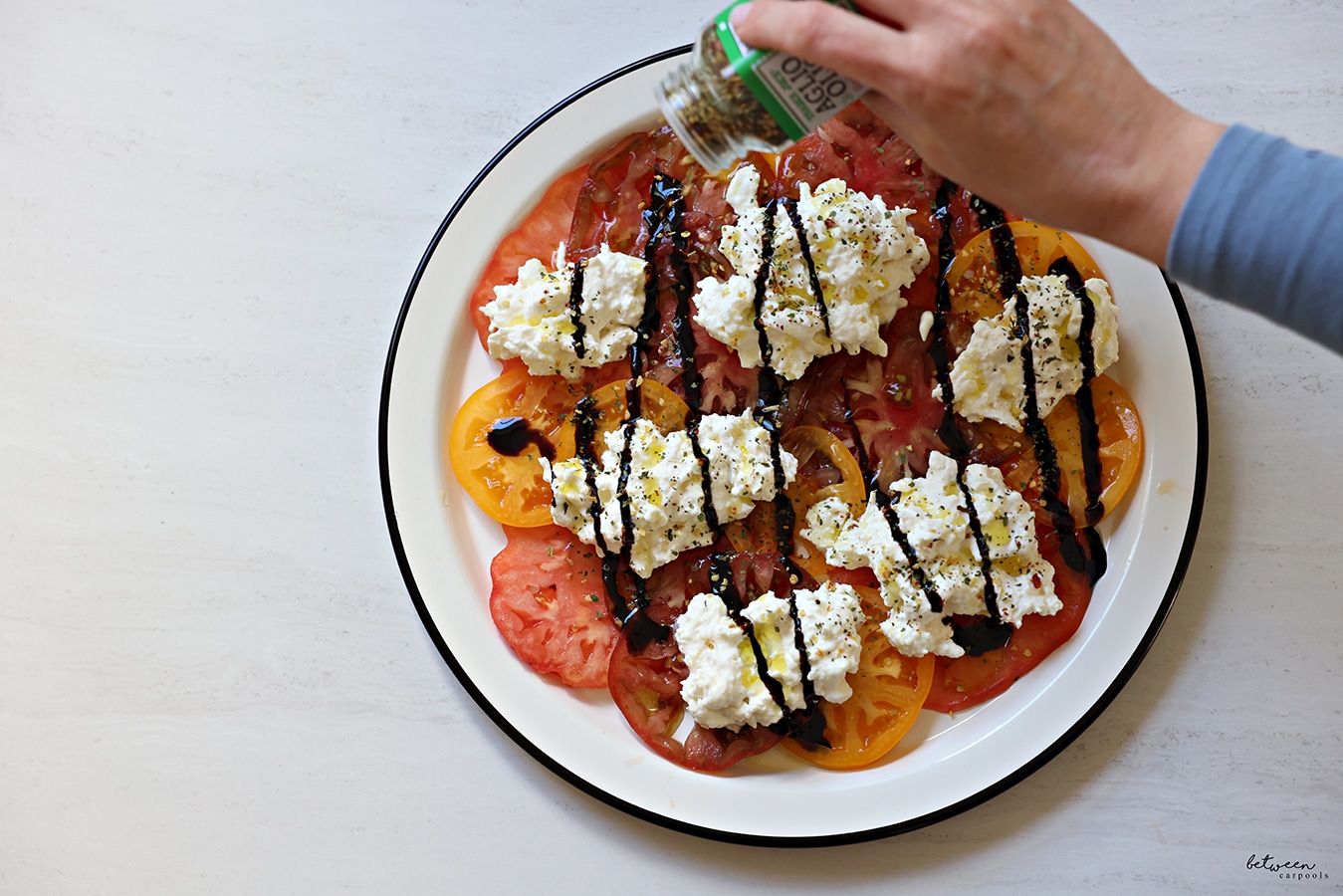 sprinkling Trader Joe’s Aglio Olio over olive oil and balsamic glaze which is over dollops of soft cheese on tomatoes - carpaccio style - on a round plate