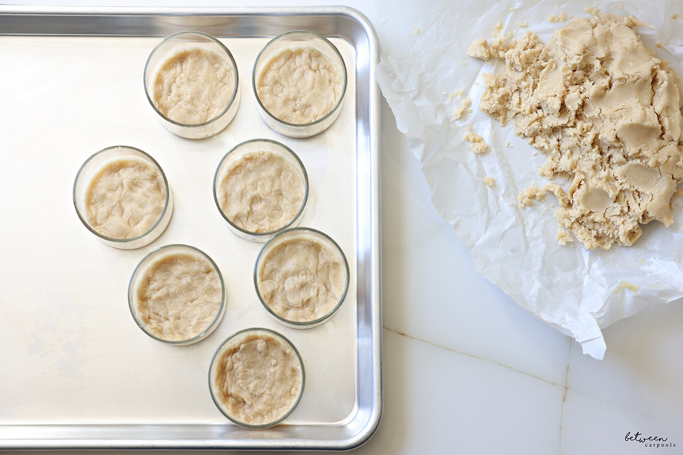 Ramekins with shortbread cookie base on a baking sheet ready for the oven. Cookie dough nearby on parchment paper