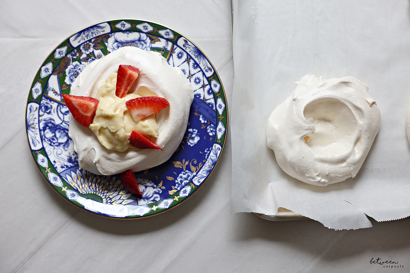 Plated pavlova with lemon curd and strawberries. Pavlova on parchment lined baking sheet nearby.