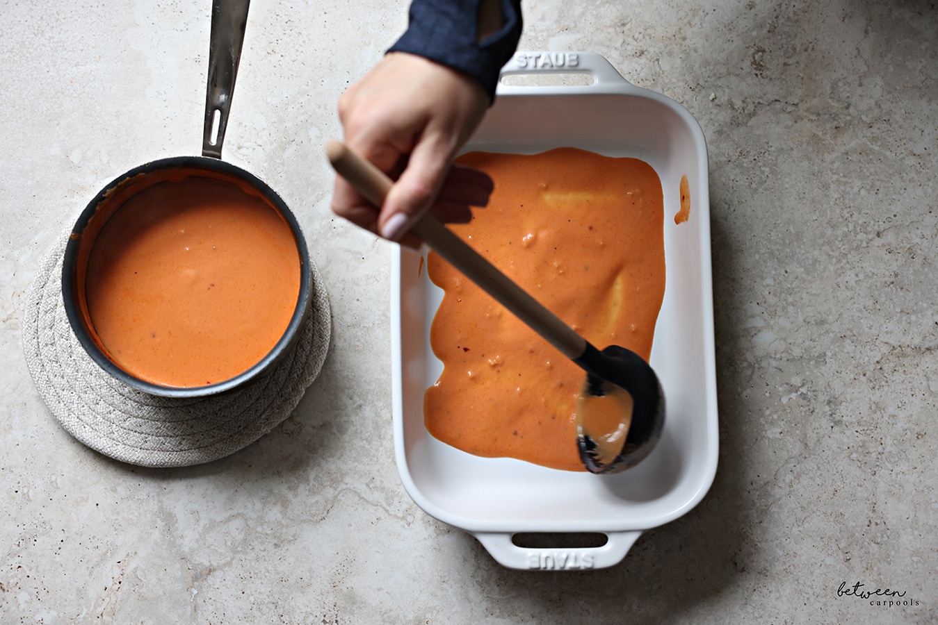 Lining the baking pan with some cream sauce using a ladle. Pot of cream sauce nearby.