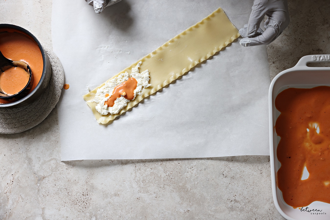 A strip of lasagna with cheese sauce topped with some tomato cream sauce, on a piece of parchment paper.