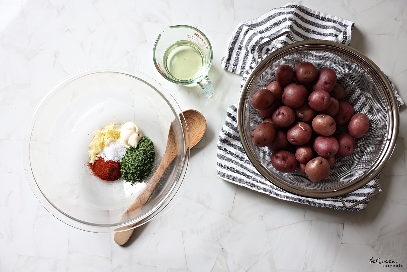 Baby red potatoes in a mesh strainer on a tea towel. Spice mixture in a glass bowl, wooden spoon nearby. Oil in a Pyrex measuring cup.