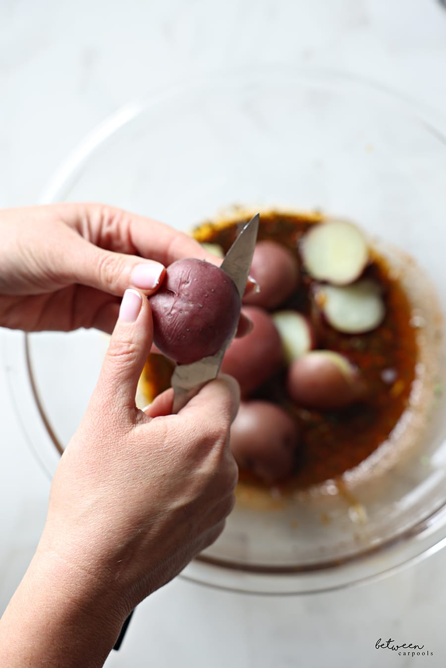 Slicing boiled potatoes in half over a glass bowl and adding them to the mixture in the bowl.