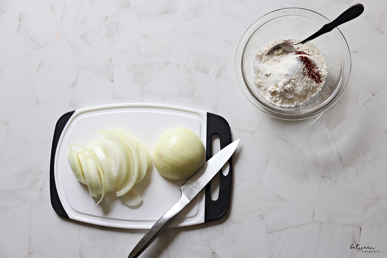 Half an onion face down on a white plastic cutting board with black handles, half cut into strips. Flour, salt, and chili powder in a glass pyrex bowl.
