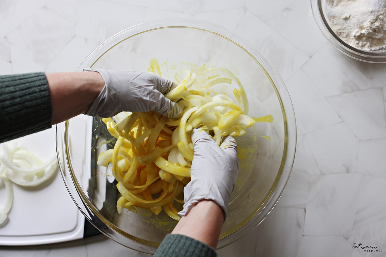 Tossing onion strips with honey, mustard, and vinegar in a pyrex bowl. Onion strips on a white plastic cutting board with black handles. Flour, salt, and chili powder in a glass pyrex bowl.