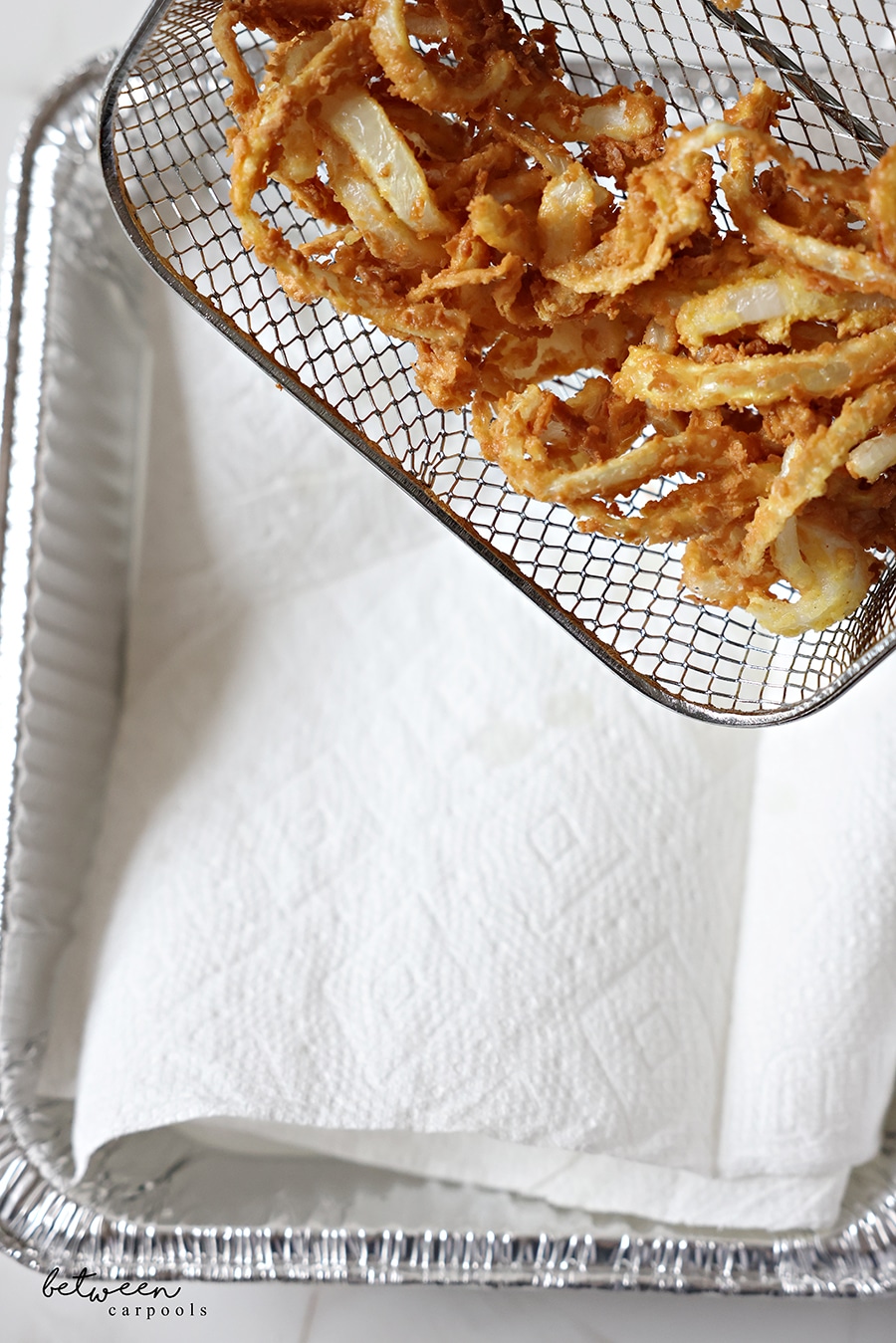 Transferring deep fried onion strings from electric deep fryer basket to a paper towel lined 9x13 aluminum pan.