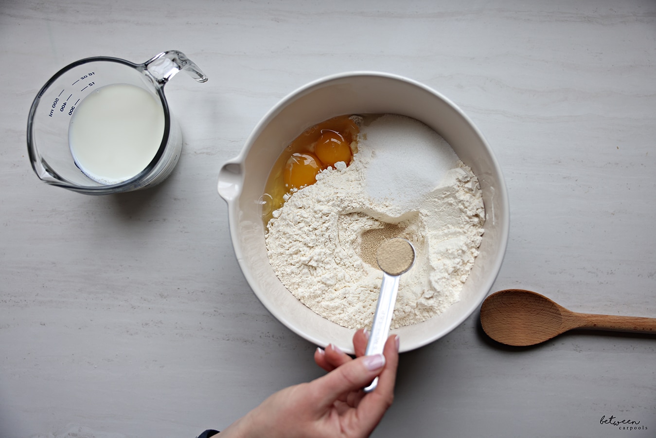 Adding dry yeast to a bowl with flour, sugar, and eggs. Milk in a pyrex glass measuring cup and a wooden spoon nearby.