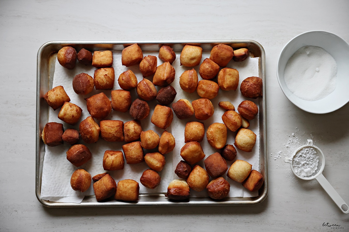 Beignets on a paper towel lined metal sheet pan. Sugar in a bowl. Confectioner's sugar in a sieve.