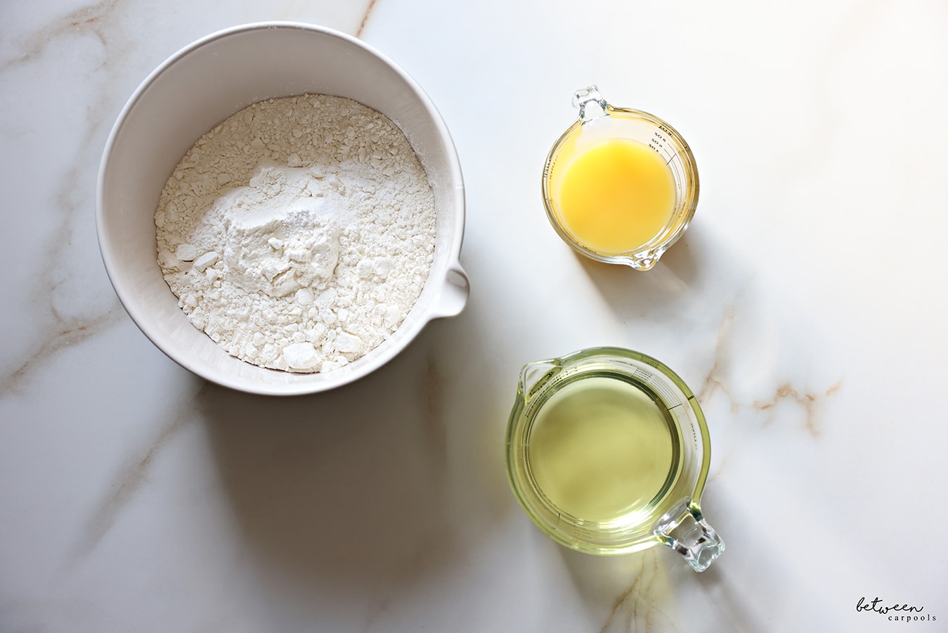 Flour in a mixing bowl, orange juice and oil in glass measuring cups.