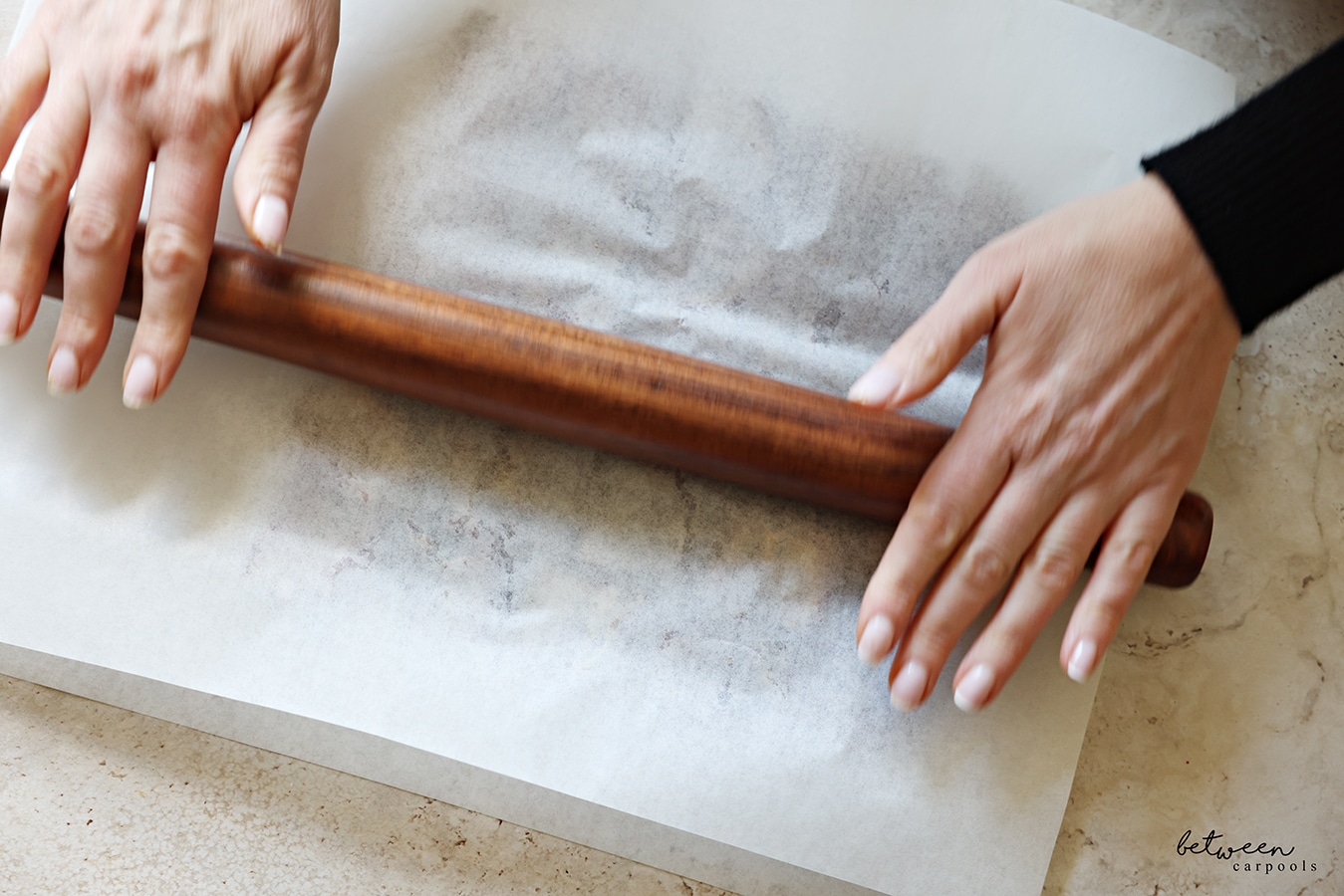 Using a rolling pin to flatten down pitted dates, laying open down, between two layers of parchment paper.