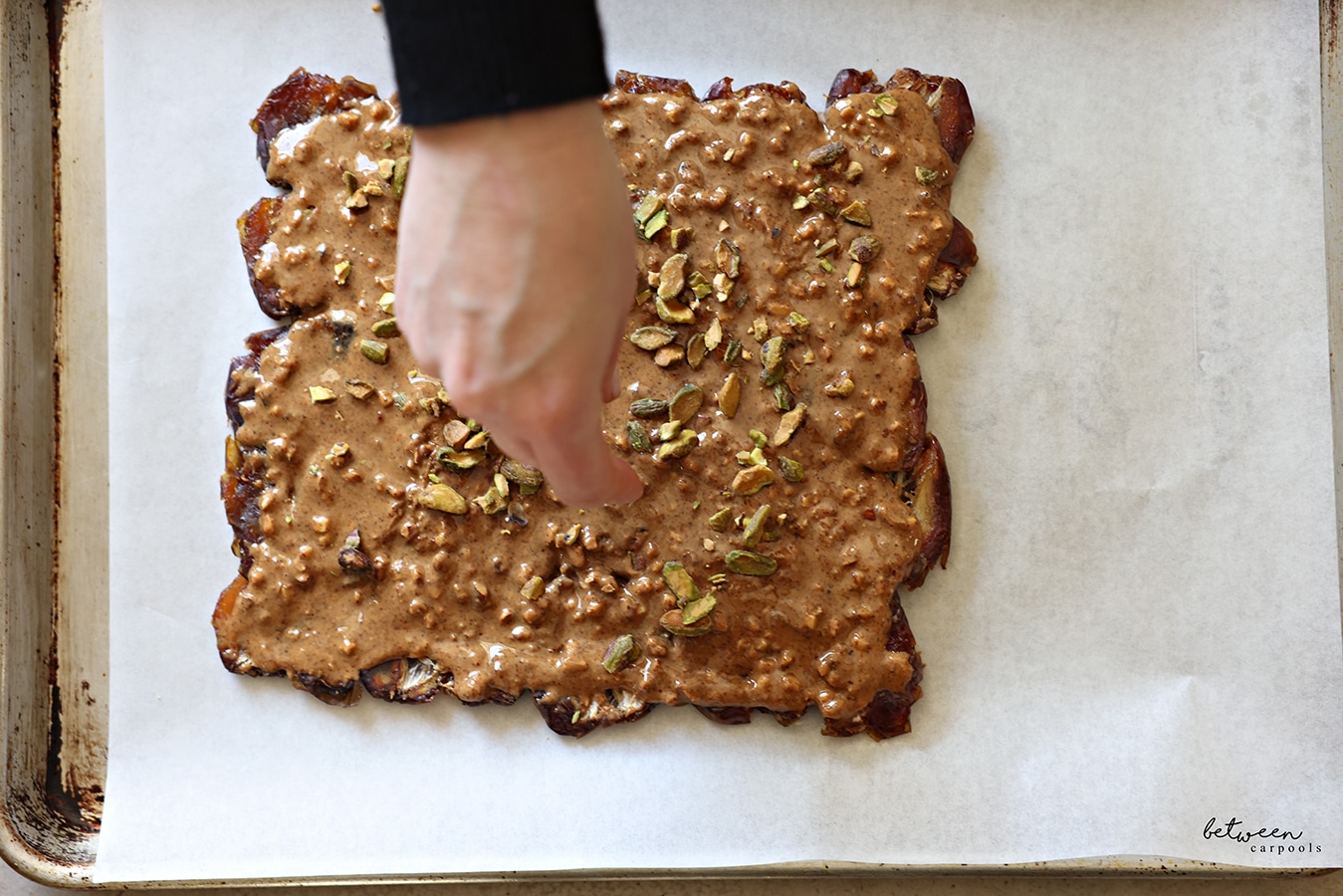 Sprinkling pistachios and walnuts over nut butter over the flattened layer of dates on a parchment lined baking sheet.