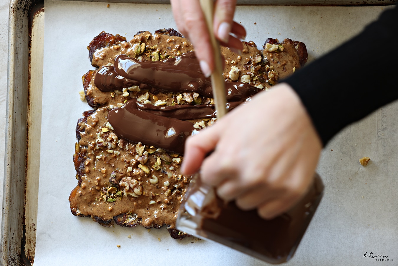 Pouring melted chocolate over the flattened layer of dates with nut butter, pistachios, and walnuts, on a parchment lined baking sheet.