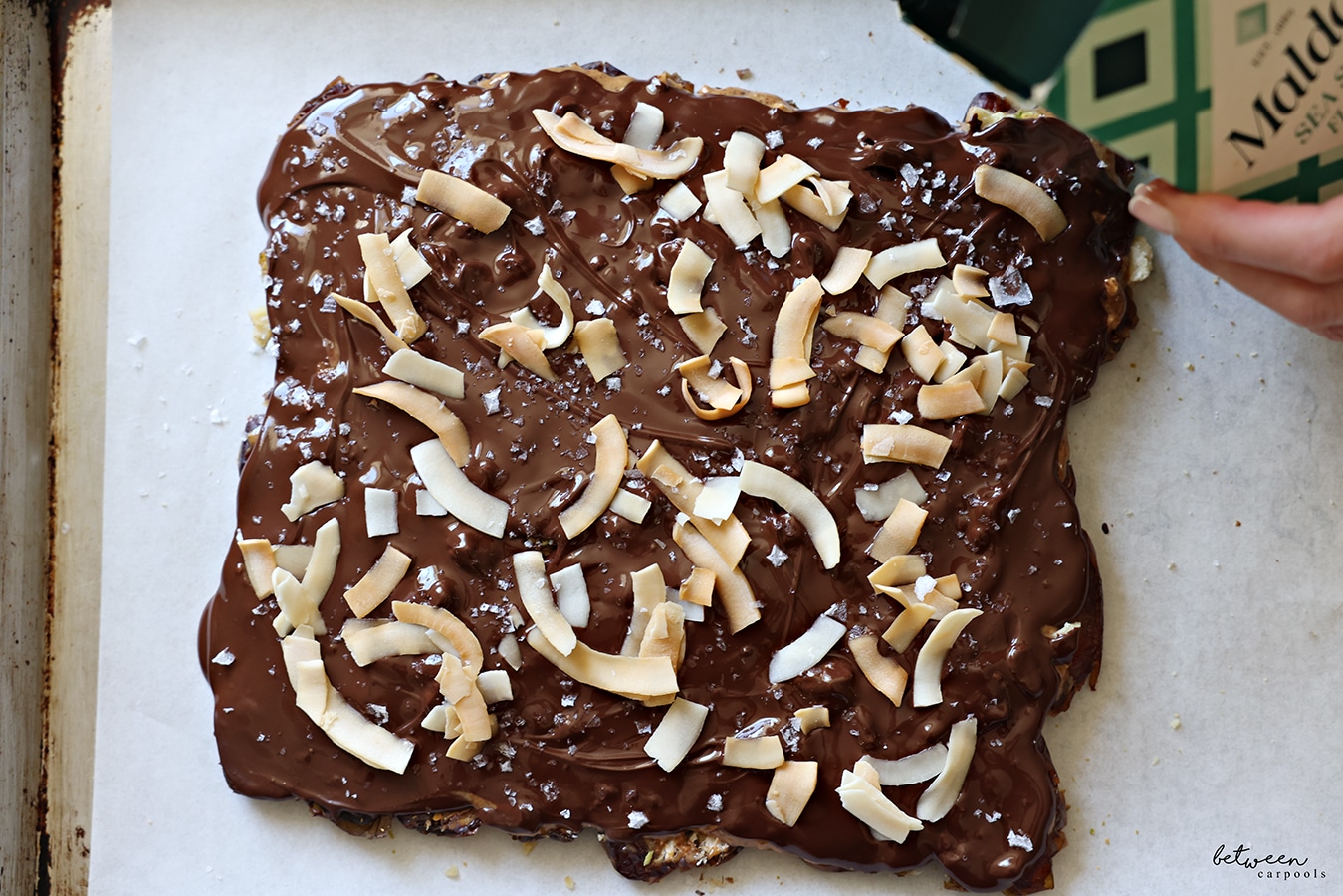 Pouring Maldon sea salt flakes over date bark - flattened layer of dates with nut butter, pistachios, walnuts, melted chocolate and coconut chips on a parchment lined baking sheet.