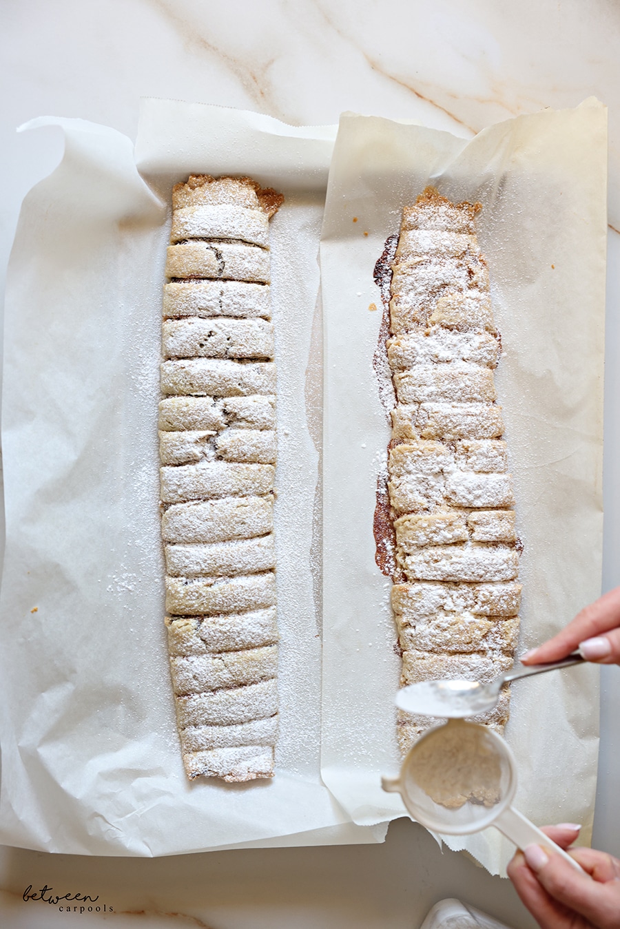 Sprinkling 2 rolls of baked date spread cookies with confectioners' sugar.