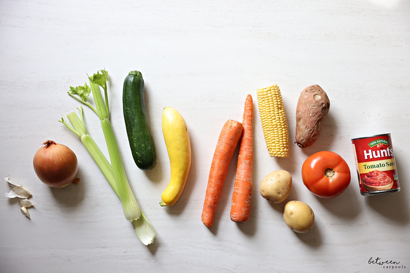 Garlic cloves, an onion, celery, a zucchini, a yellow squash, two carrots, an ear of corn, a sweet potato, two Yukon Gold potatoes, a beef tomato and a can of Hunts Tomato Sauce.