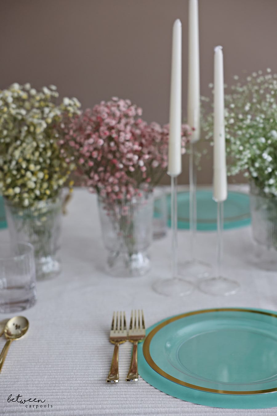 Closeup of table set with a cloth, paper placemats, plates, flatware, candles and colored baby's breath in vases.