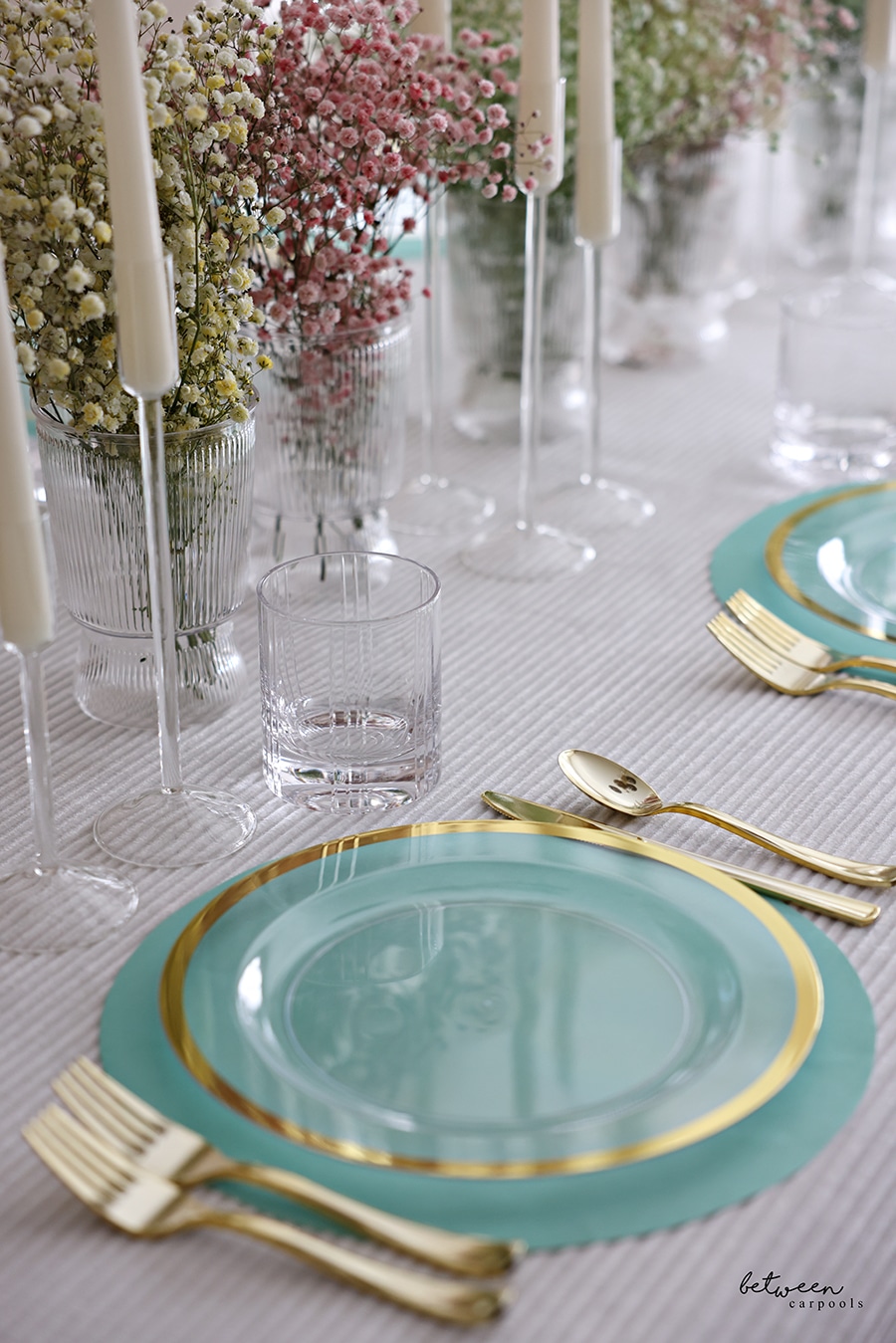 Closeup of table set with a cloth, paper placemats, plates, flatware, candles and colored baby's breath in vases.