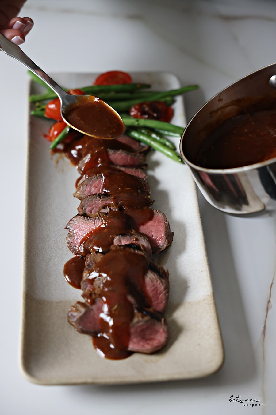 Pouring boiled marinade over sliced oyster steak, sous vide and grilled, on a serving tray with green beans and tomatoes.