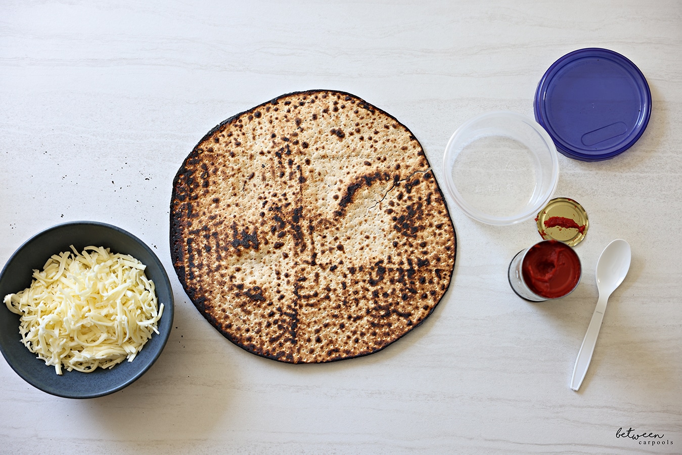 Bowl with shredded cheese, one round Shmurah Matzah, an open 16 oz twist top storage container, an open 6 oz can of tomato paste, and a plastic spoon.